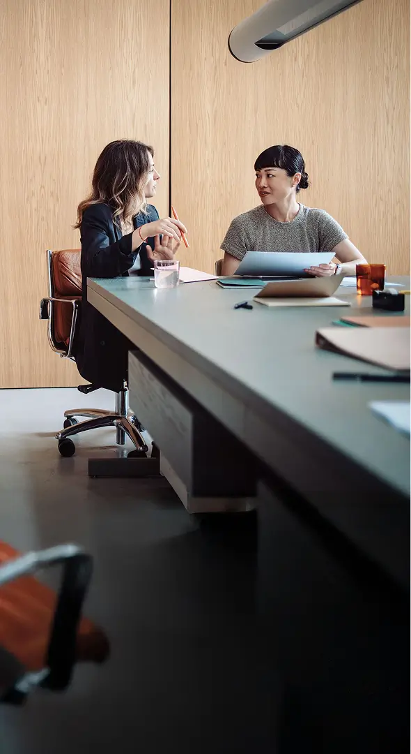 Two professionals sit at a modern boardroom table in conversation, reviewing documents with a laptop and notepads between them.
