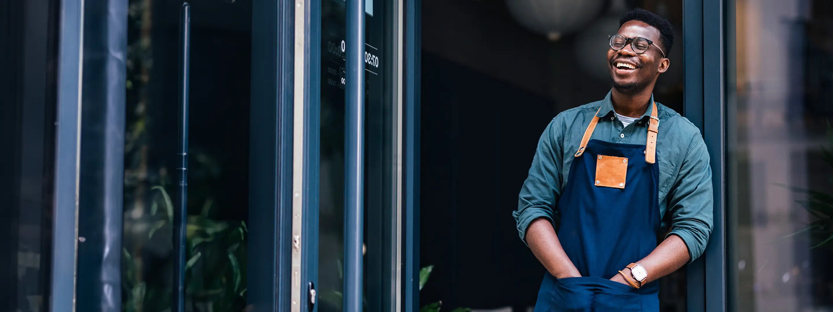 A smiling business owner stands in the doorway of their shop, wearing an apron and looking relaxed and welcoming.