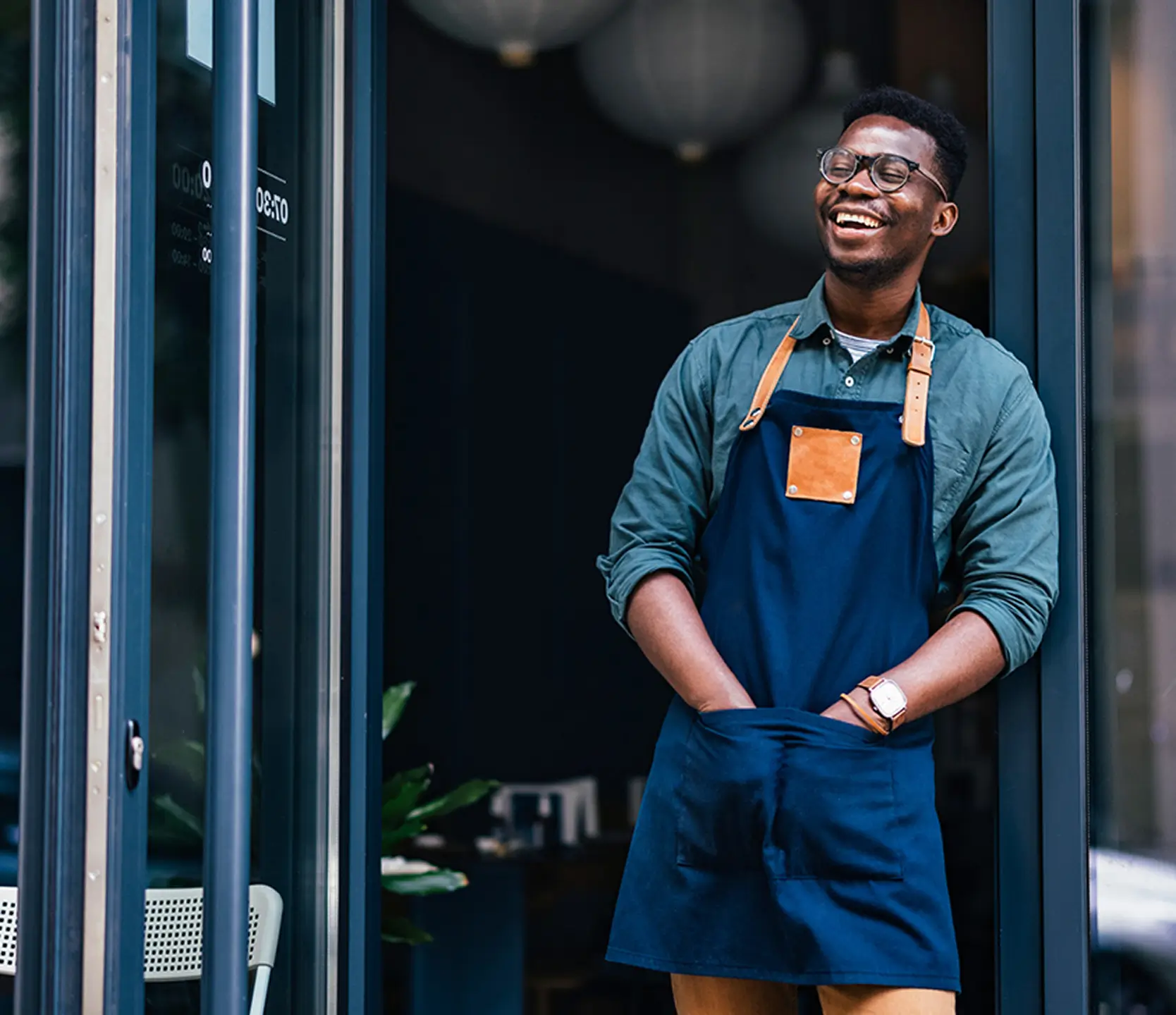 A smiling business owner stands in the doorway of their shop, wearing an apron and looking relaxed and welcoming.