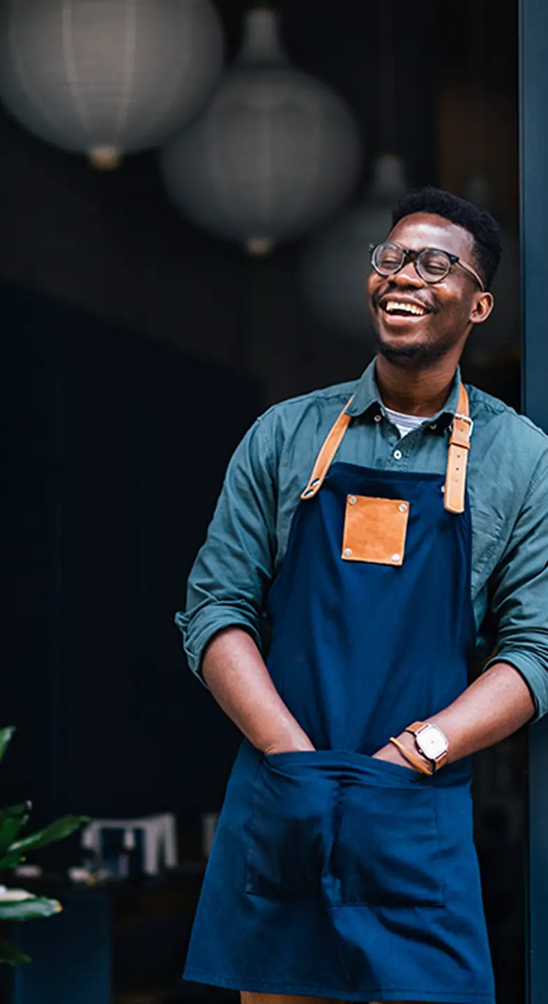 A smiling business owner stands in the doorway of their shop, wearing an apron and looking relaxed and welcoming.