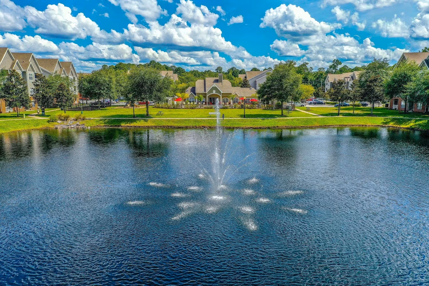 Wide view of the community lake with a fountain and the clubhouse in the distance, set against a sunny sky at The Finley in Jacksonville, FL.