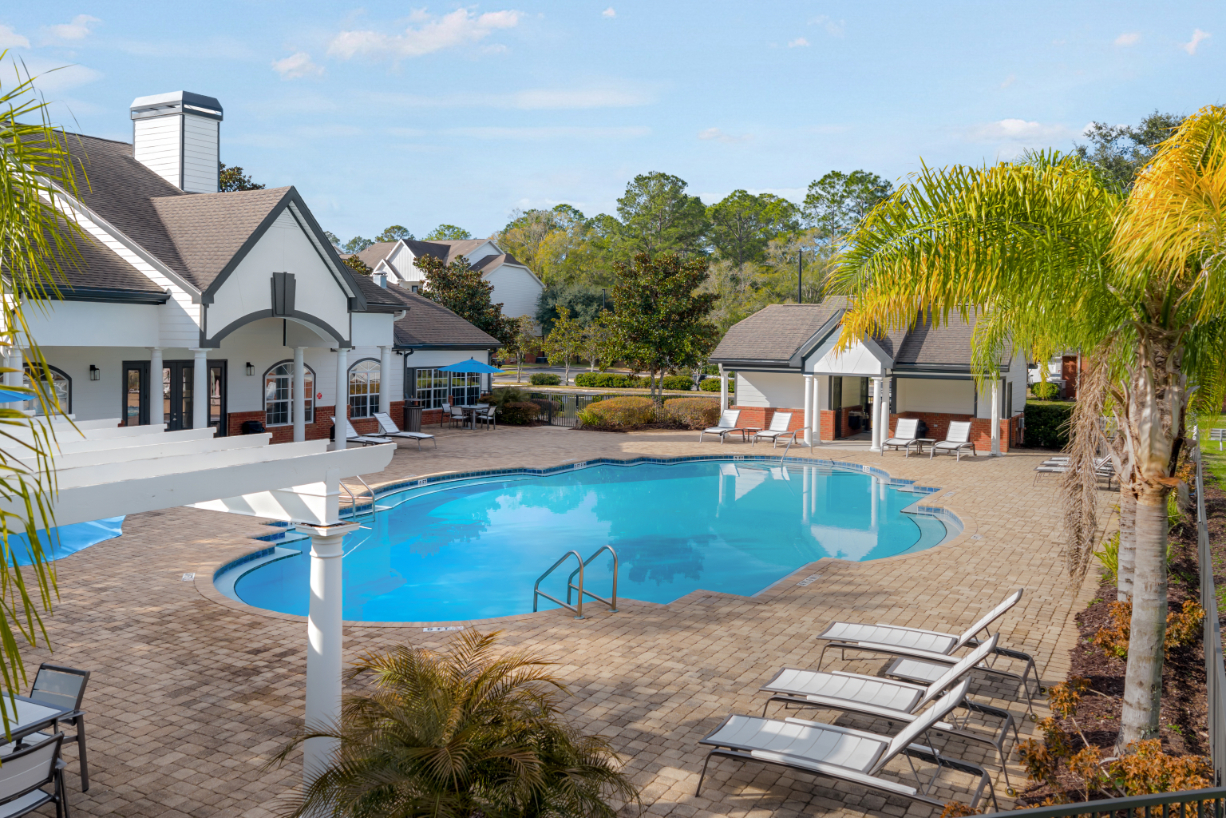 Community swimming pool with a wide brick deck, surrounded by palm trees and apartment buildings, offering a tropical feel at The Finley in Jacksonville, FL.