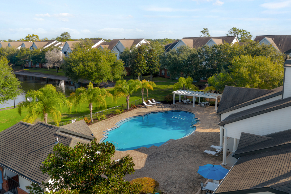 Drone shot view of Community swimming pool with a wide brick deck, surrounded by palm trees and apartment buildings, offering a tropical feel at The Finley in Jacksonville, FL.