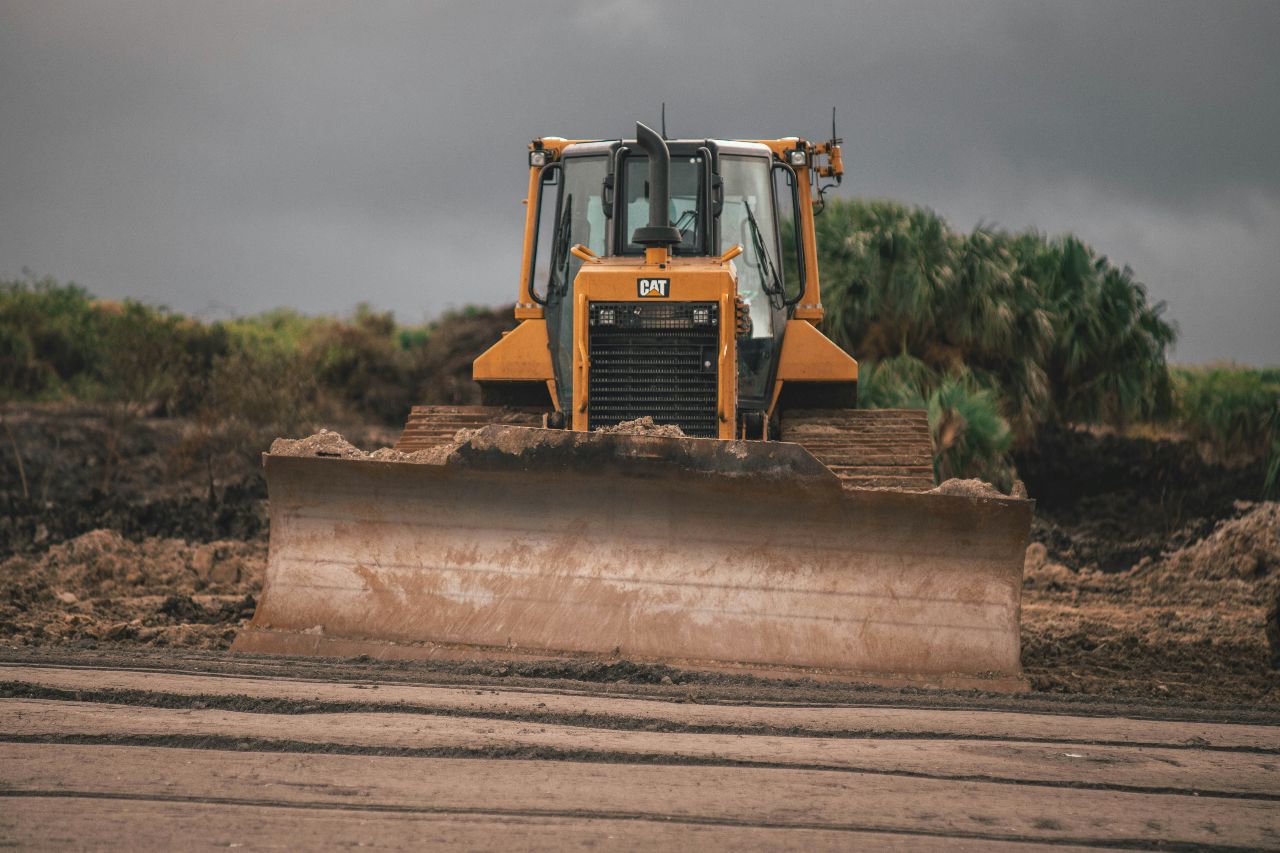 Yellow bulldozer pushing dirt on construction site under cloudy sky, large blade lowered, surrounded by muddy terrain and vegetation.