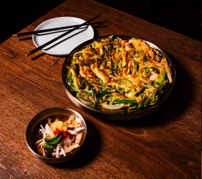 Korean vegetable pancake served on a round black plate with a small bowl of dipping sauce containing sliced vegetables and chili, placed on a wooden table with a white plate and black chopsticks beside it.
