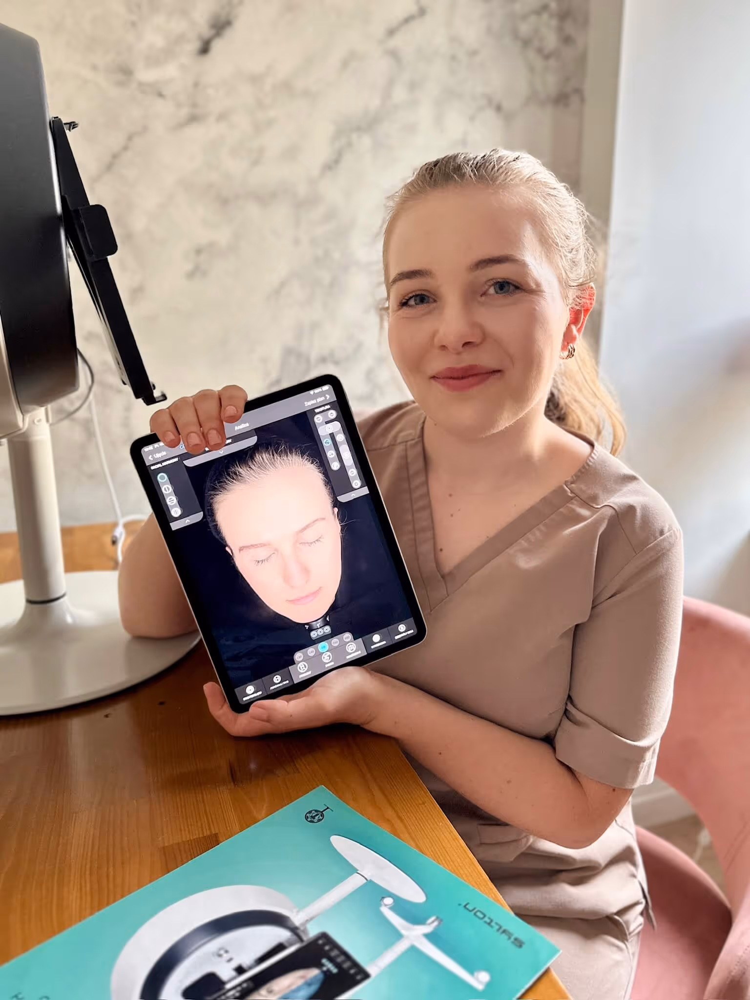 Smiling woman in beige scrubs holding a tablet displaying a 3D facial scan with closed eyes.