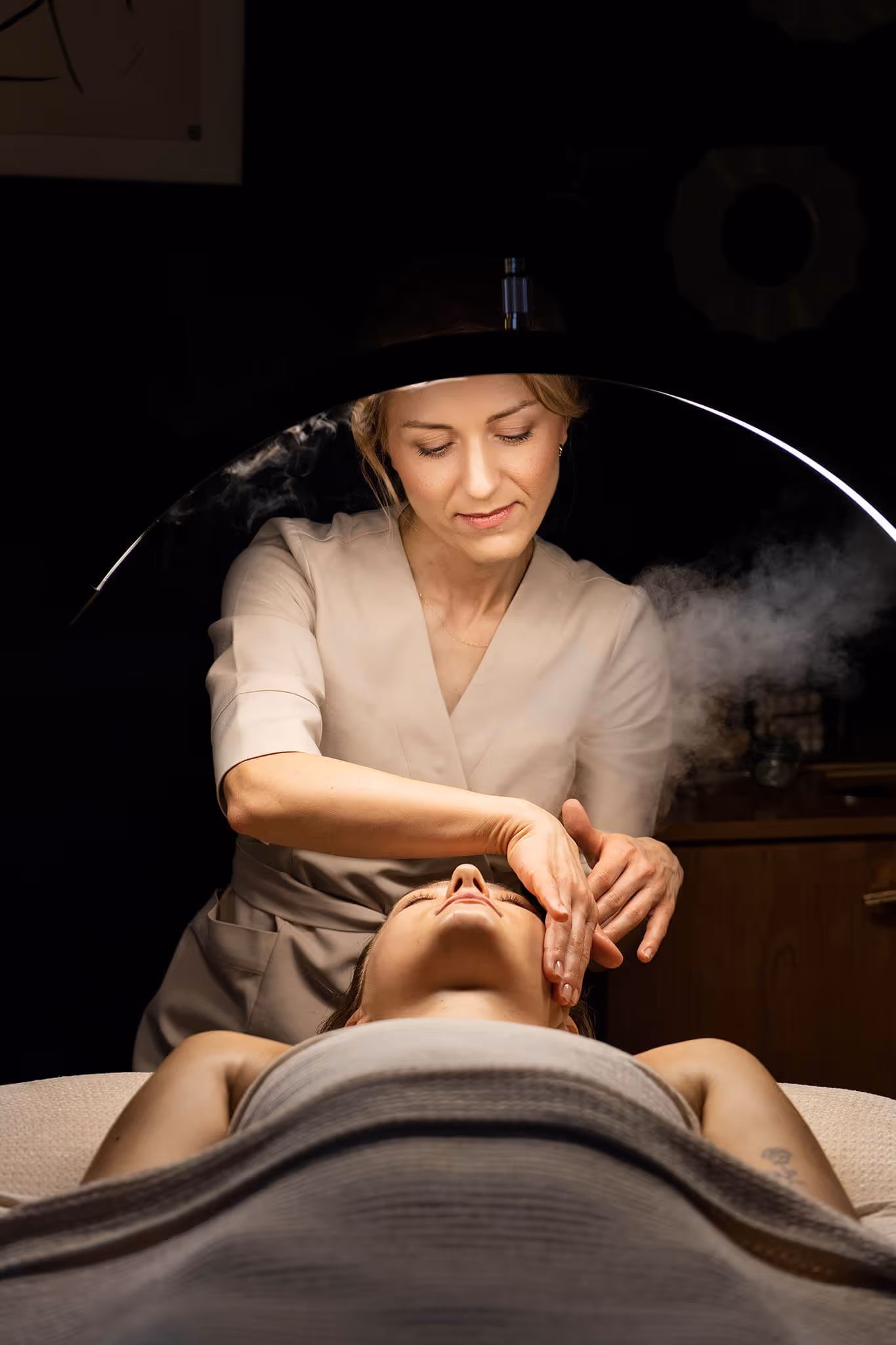 Spa therapist gently massaging a woman's face under a curved lamp with steam in a dimly lit room.