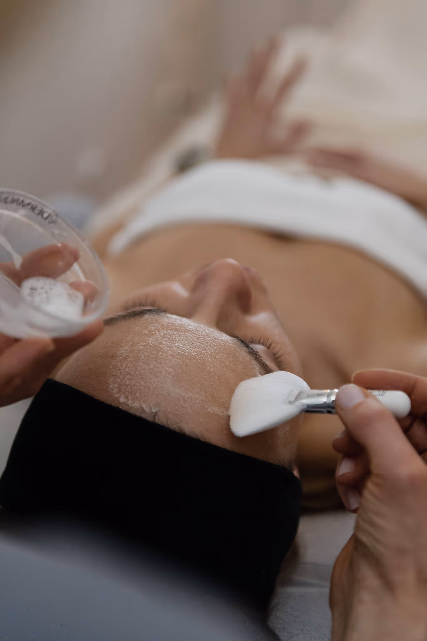 Person lying down with eyes closed while a skincare professional applies a facial mask with a brush.
