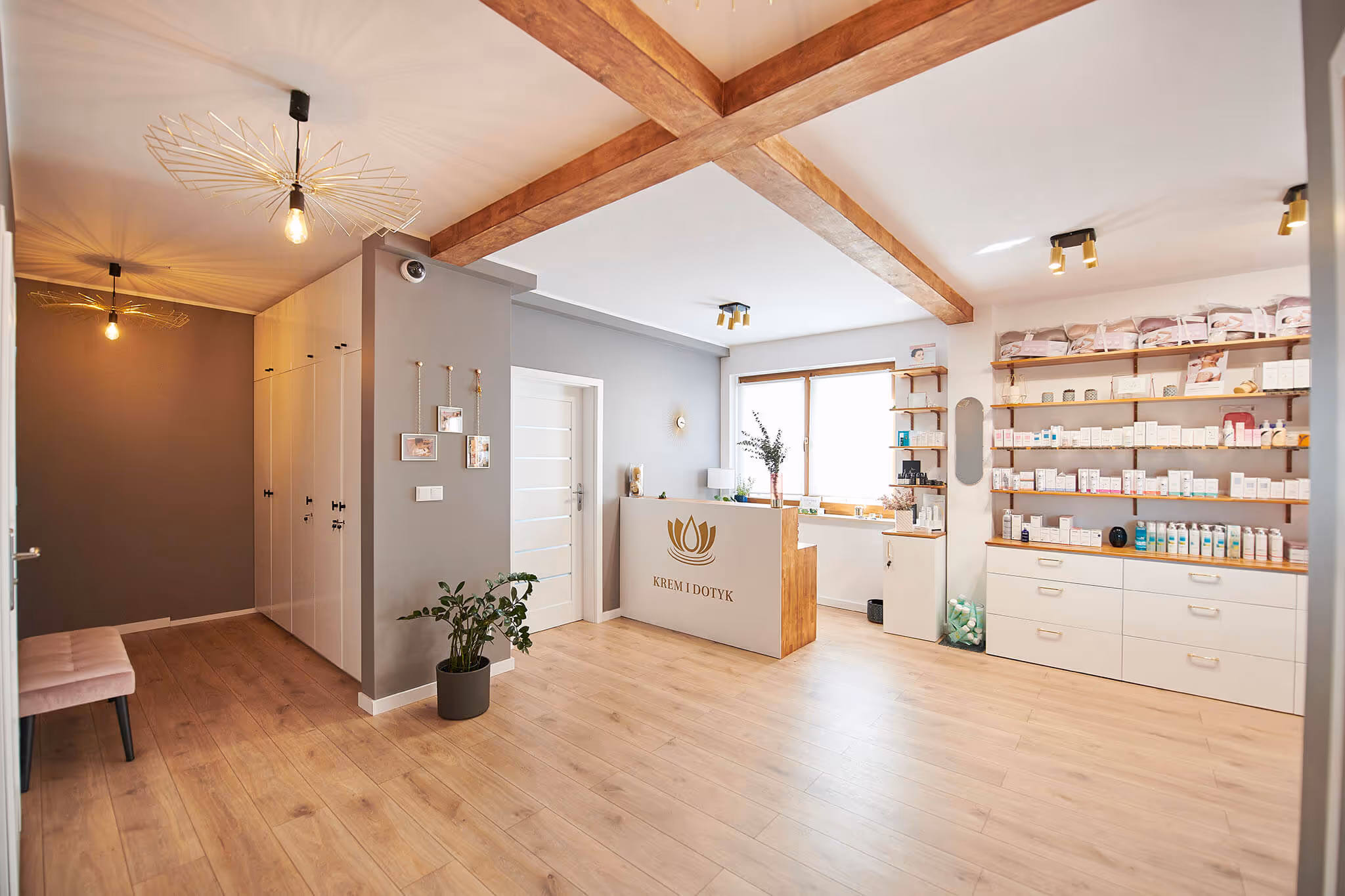 Interior of a bright spa or beauty salon reception area with wooden floor, wooden ceiling beams, modern pendant lights, a white reception desk with a lotus logo and shelves stocked with skincare products.