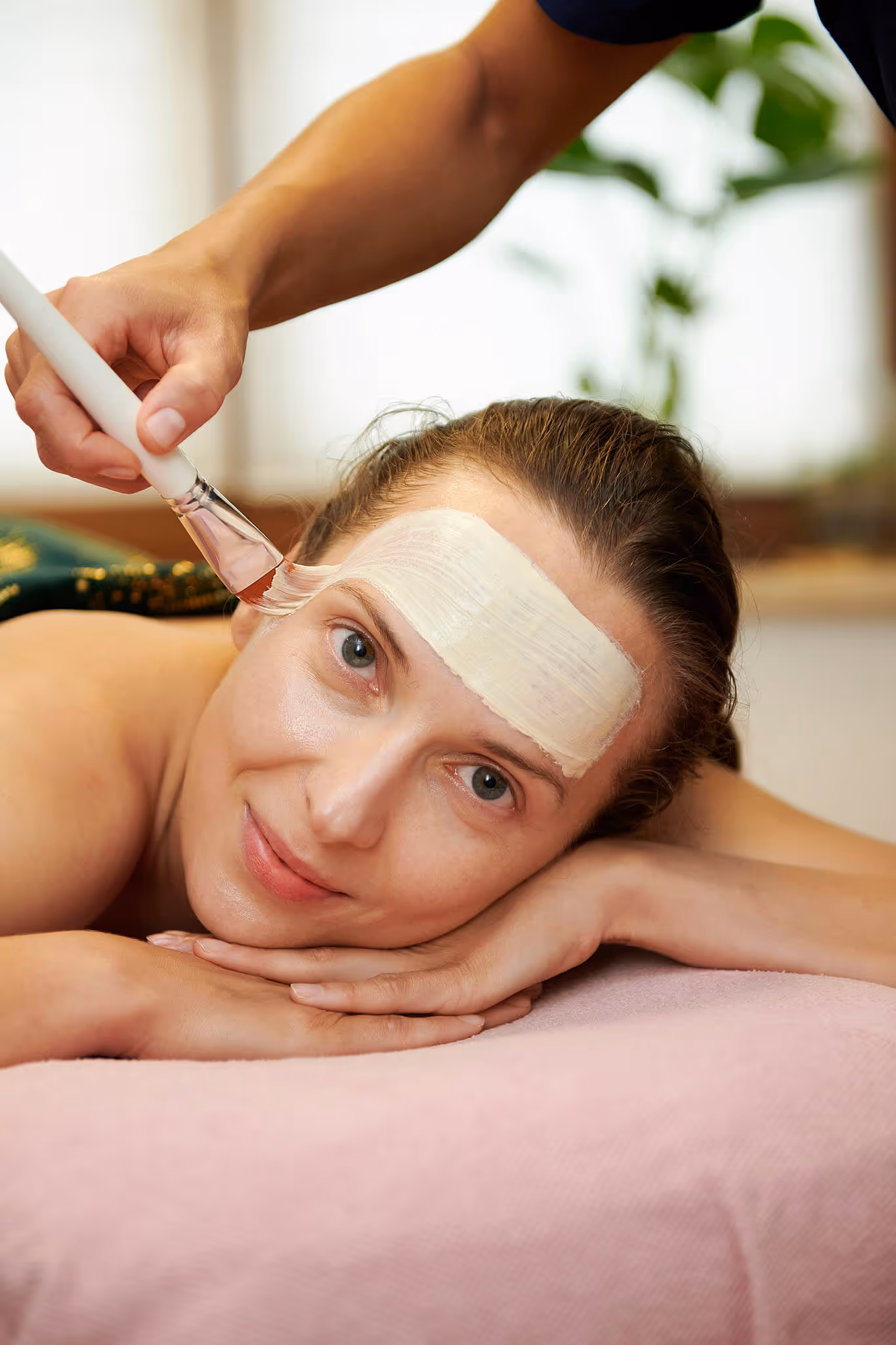 Woman lying face down on a pink towel with a white facial mask being applied to her forehead with a brush.