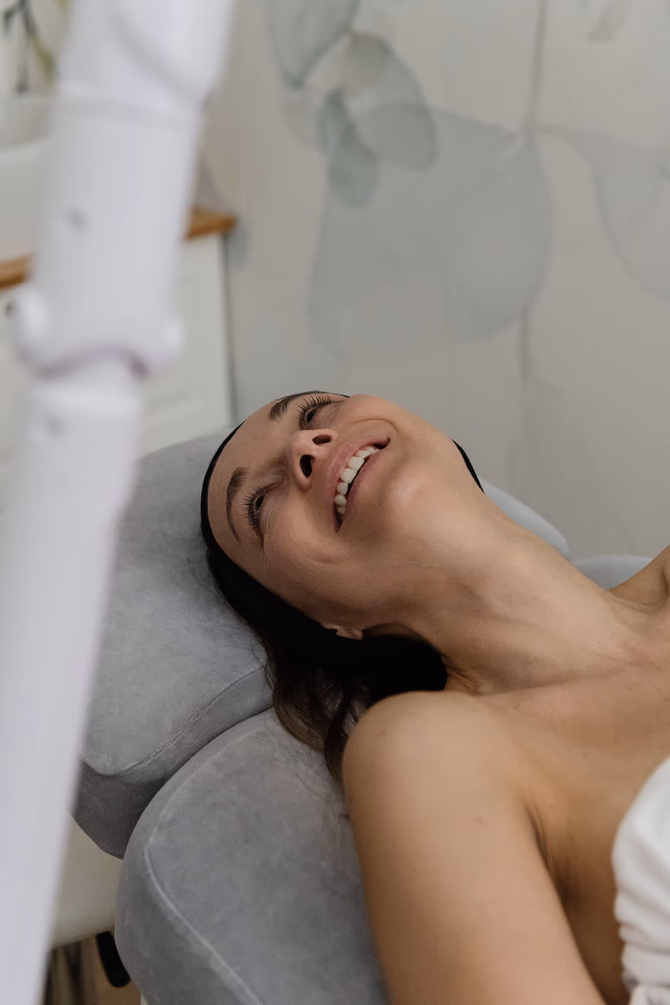 Smiling woman lying on a gray cushioned surface, looking up in a spa or treatment setting.