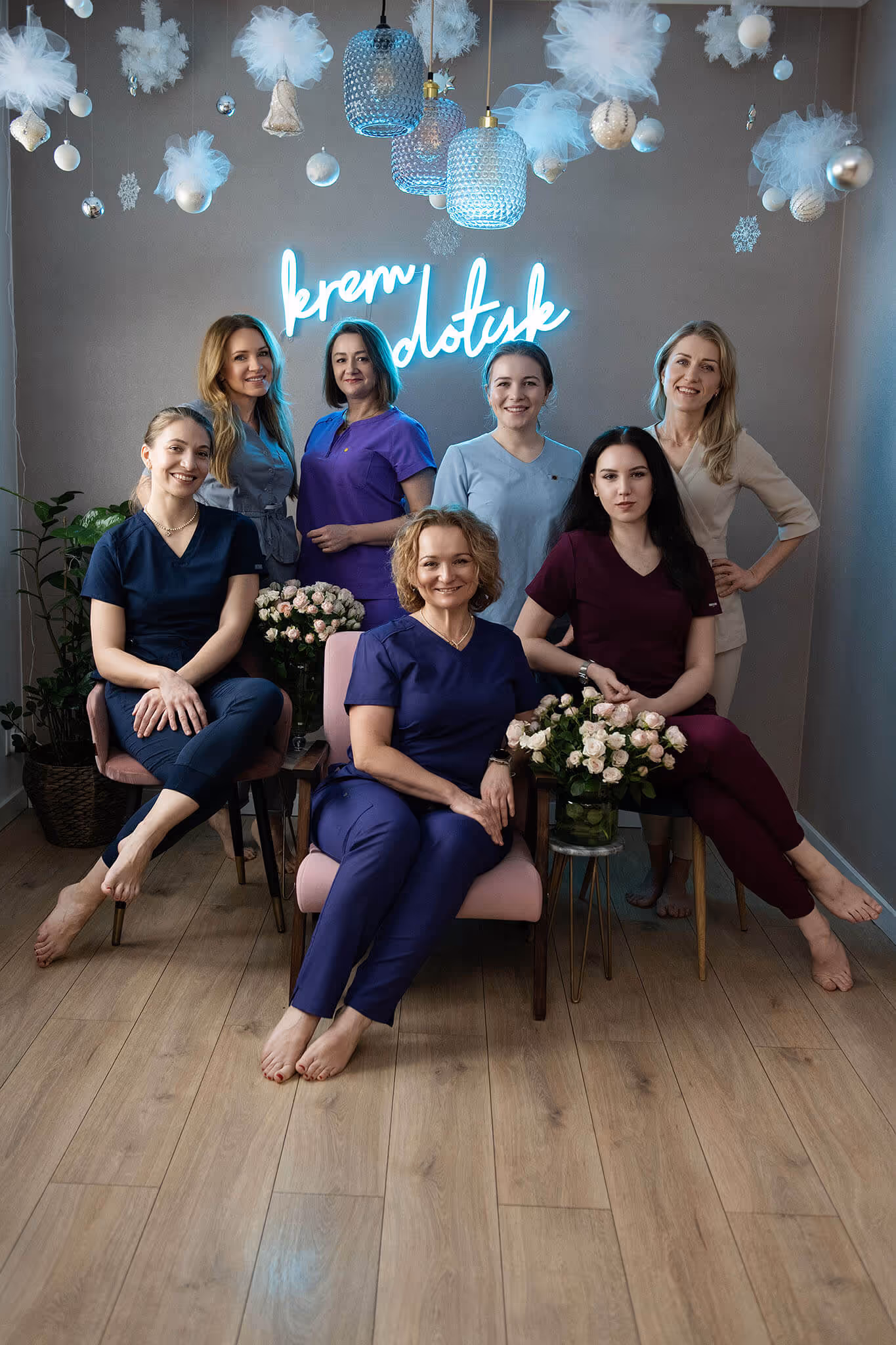 Seven women in colorful medical scrubs sitting and standing in a decorated room with hanging ornaments and a neon sign on the wall.