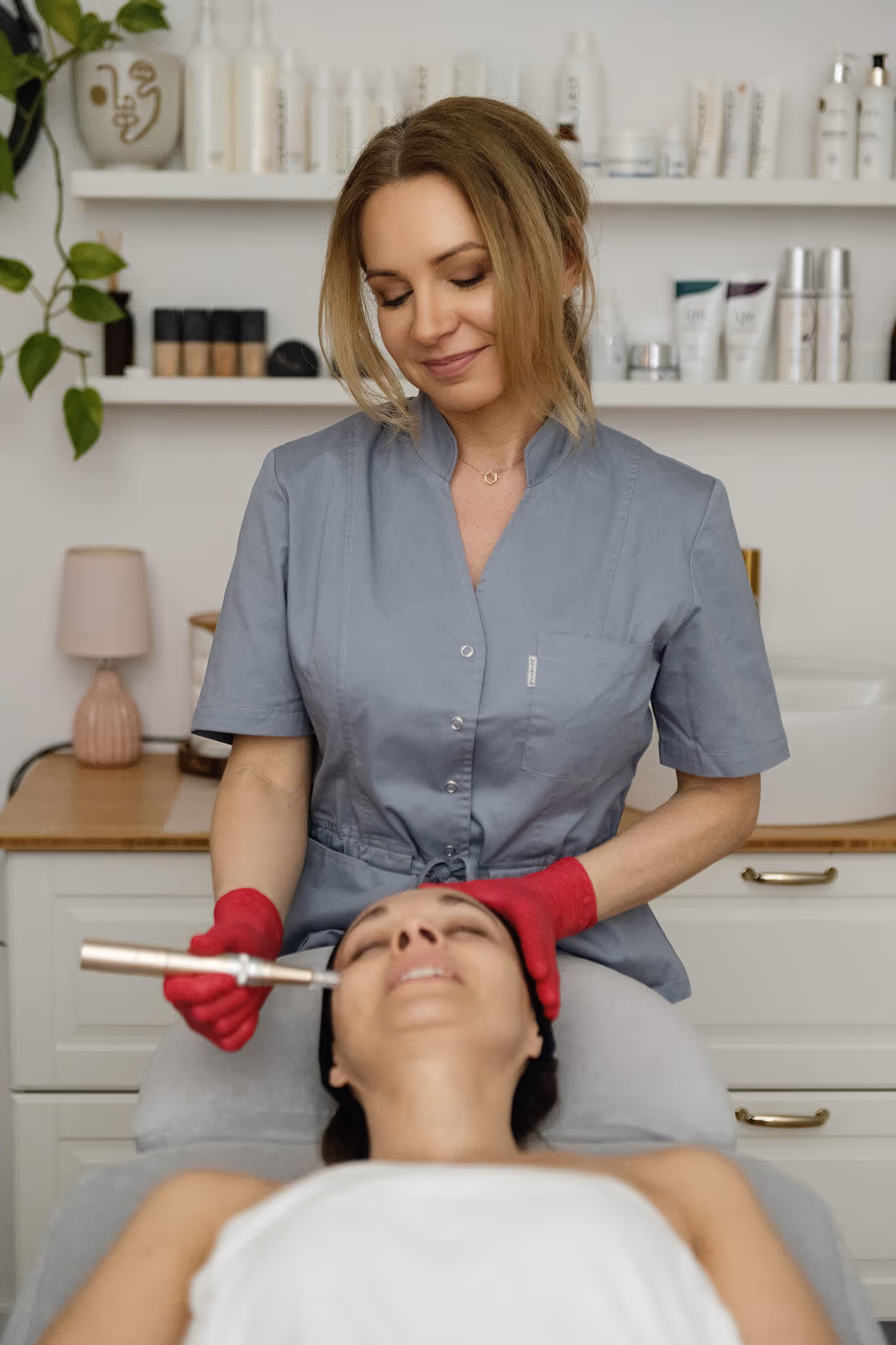 Woman cosmetologist wearing red gloves performing a facial treatment on a client lying down with eyes closed in a spa setting.