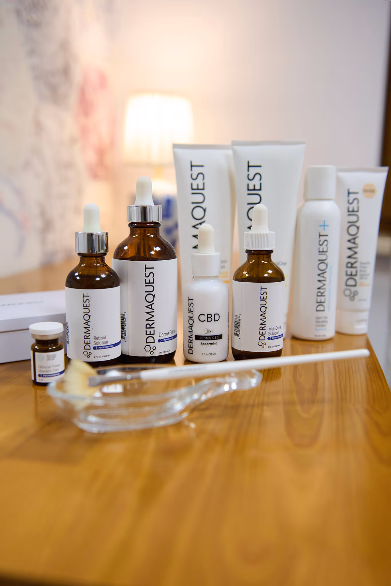 Various DermaQuest skincare products arranged on a wooden surface with a makeup brush and glass dish in front.
