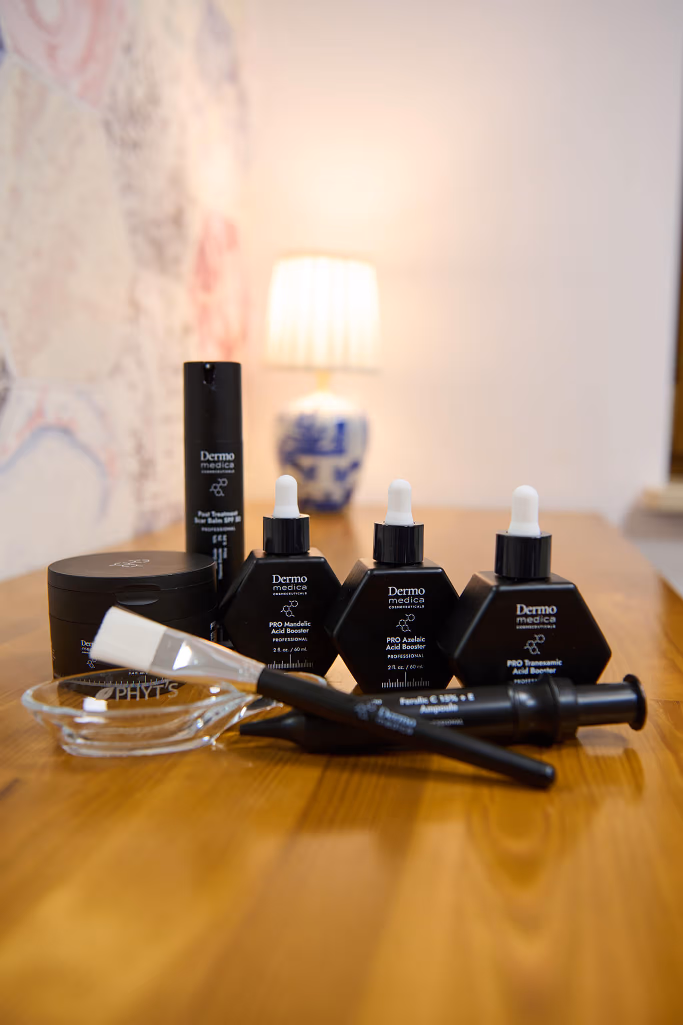 Dermo medica skincare products including acid boosters, scar balm, and brush tools arranged on a wooden surface with a lit table lamp in the background.