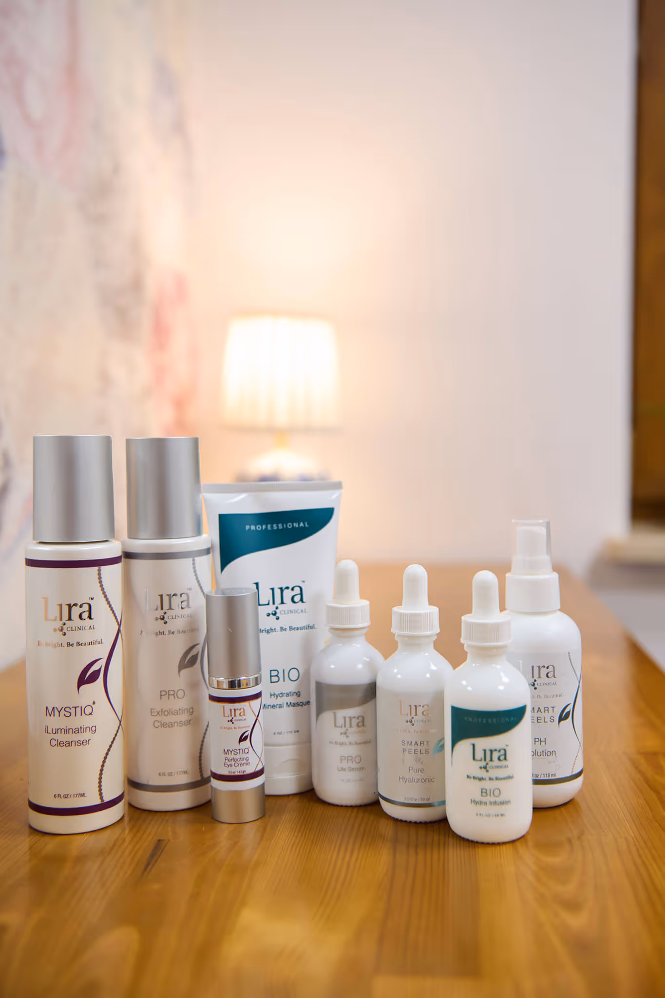 Eight Lira Clinical skincare products including cleansers, serums, eye cream, masks, and solutions arranged on a wooden surface with a blurred lamp in the background.