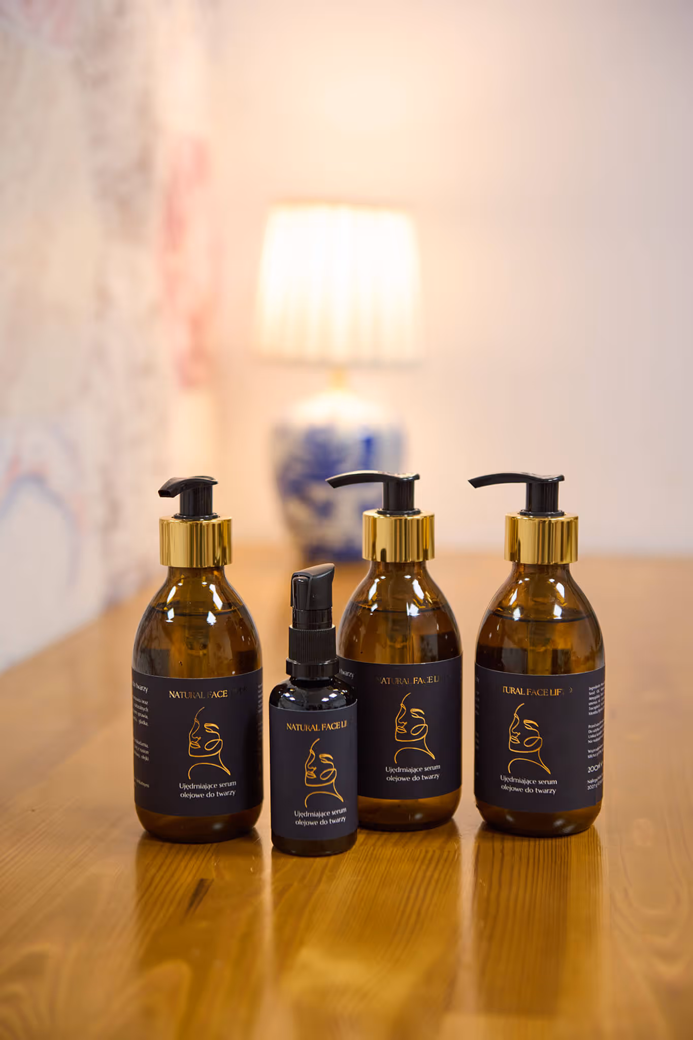 Four amber-colored cosmetic bottles with black and gold pump tops labeled 'Natural Face Lift' and placed on a wooden table with a blurred lamp in the background.
