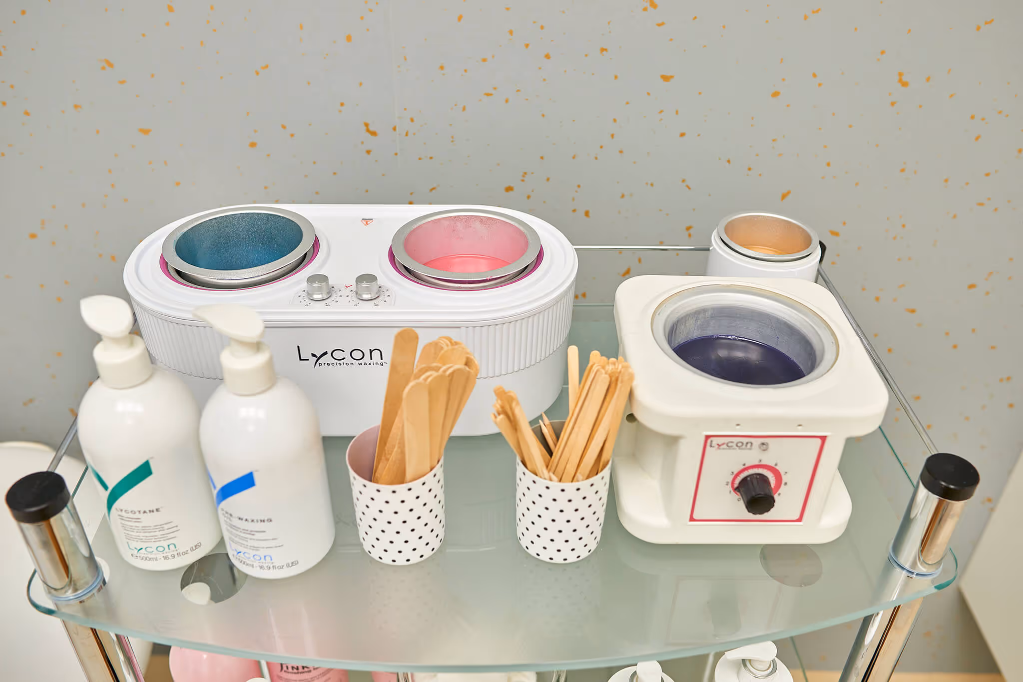 Glass shelf with two wax warmers, bottles of Lycon waxing products, and containers holding wooden applicator sticks.