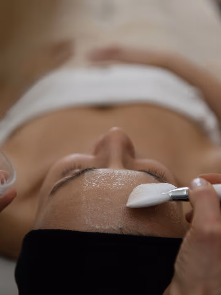Woman lying down with eyes closed receiving a facial treatment using a brush on her forehead.