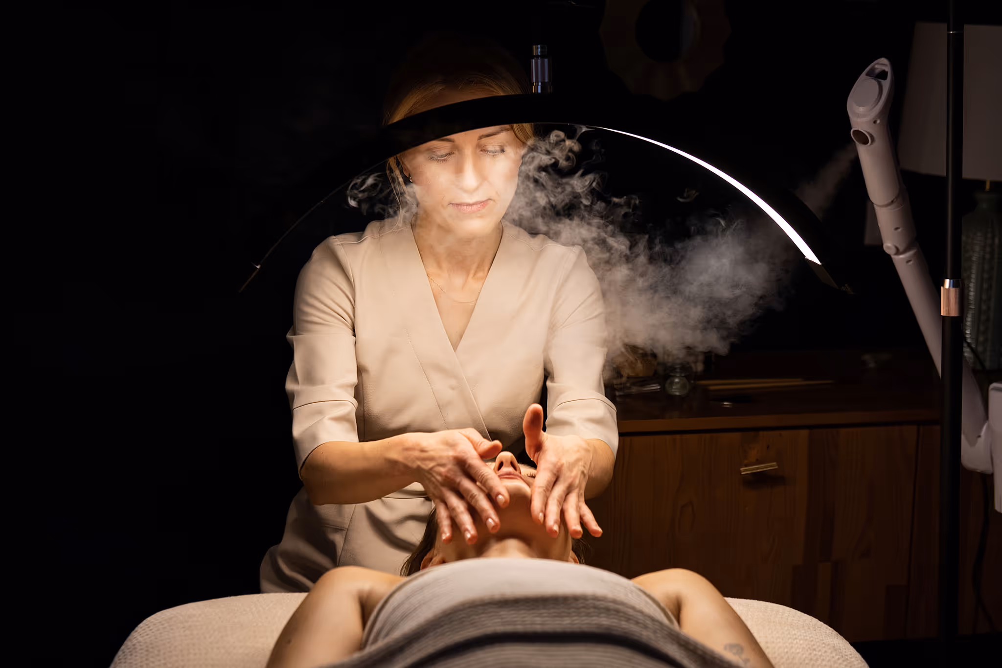 Facialist massaging a woman's face under a circular LED lamp with facial steam in a spa room.