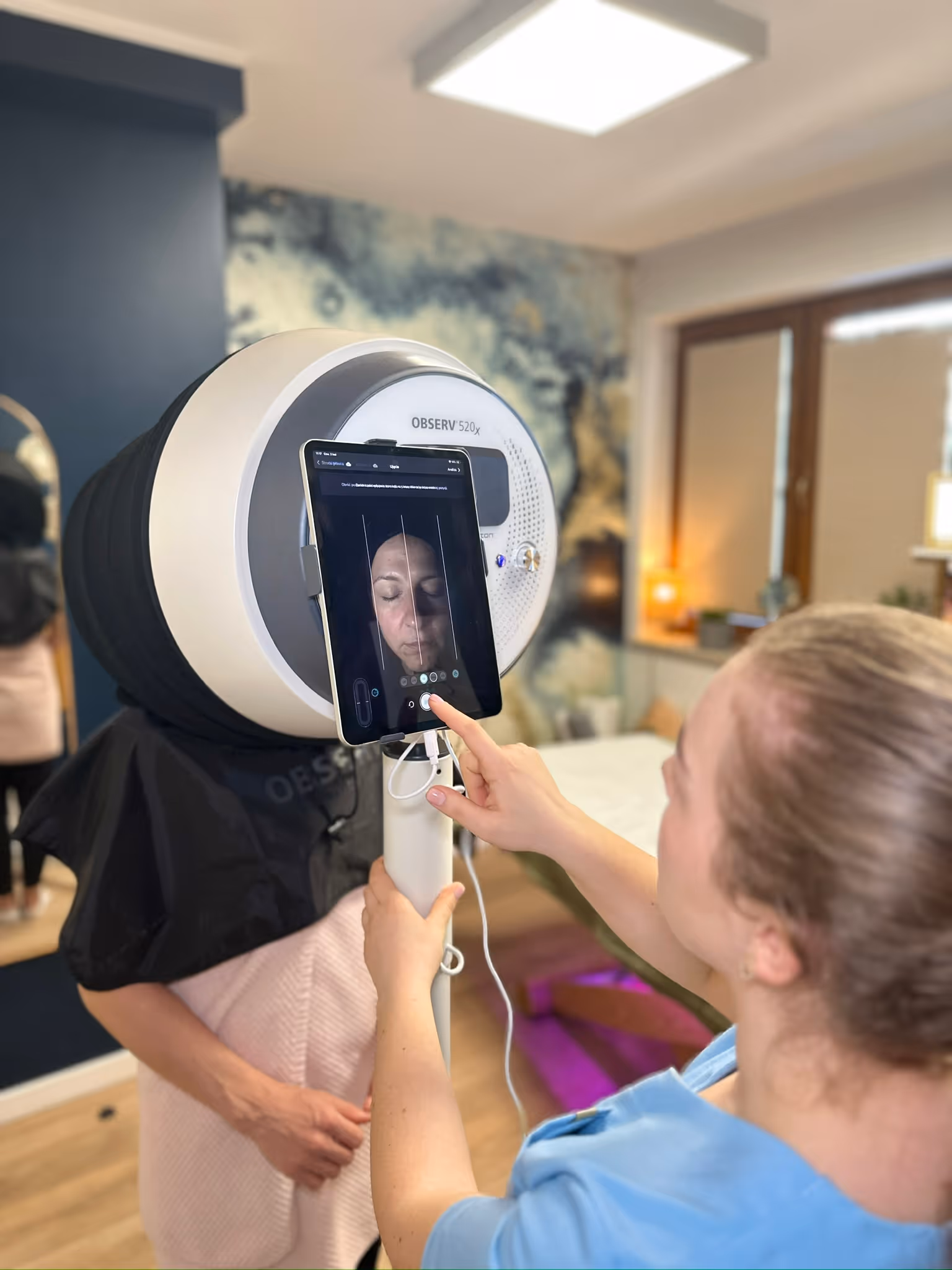 Woman operating Observ 520x skin analysis device on another woman's face in a clinic.