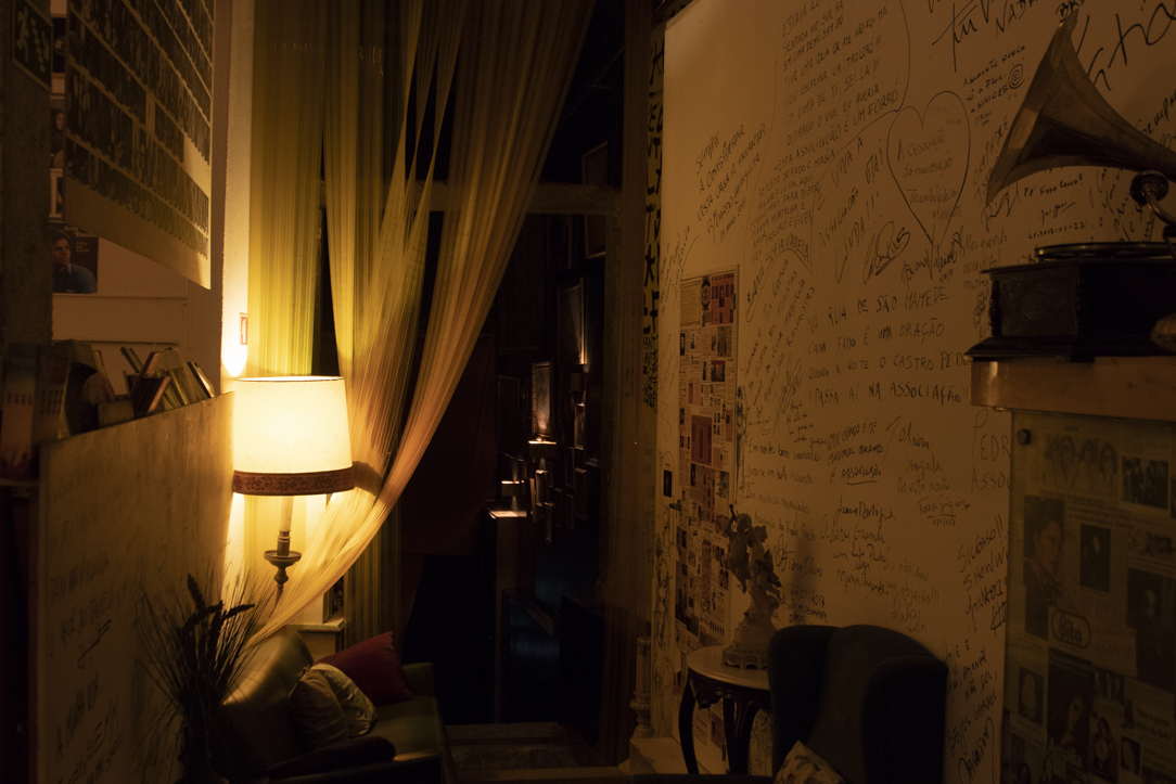 Dimly lit cozy room with a glowing lamp, sheer curtains, a couch with pillows, and walls covered in handwritten notes and photos.