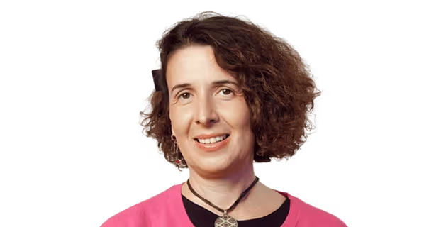 Smiling woman with curly brown hair wearing a pink top and a black choker necklace against a black background.