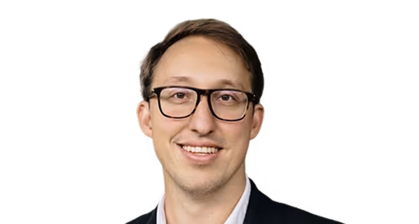 Smiling man with short brown hair wearing black glasses and a dark blazer over a white shirt.