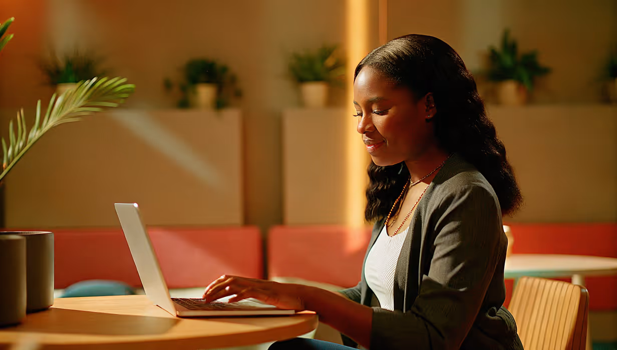 Woman sitting at a table typing on a laptop in a warmly lit indoor space with plants in the background.