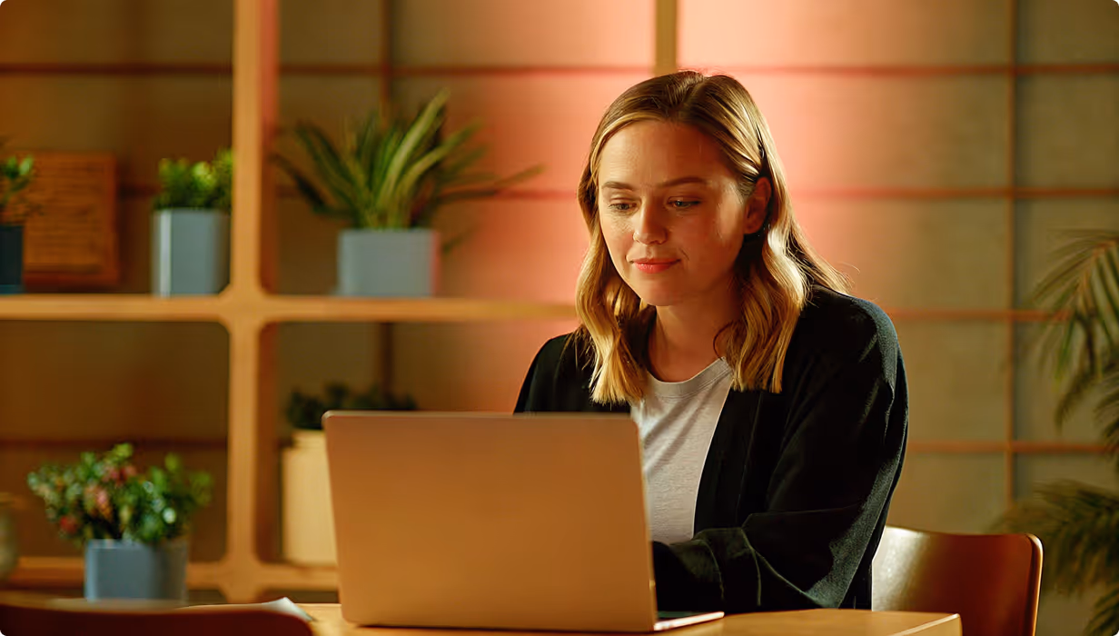 Woman with shoulder-length blond hair working on a laptop at a wooden table with potted plants on shelves in the background.