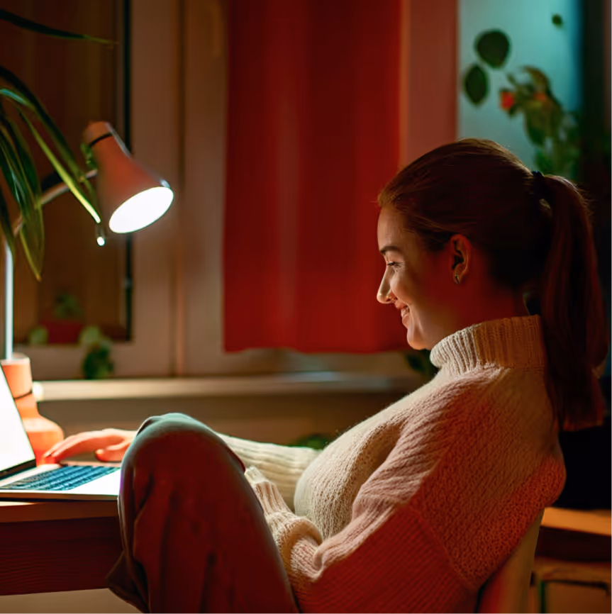 Young woman smiling while using a laptop in a warmly lit room with a desk lamp and plants.