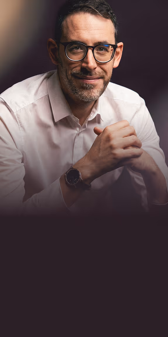 Smiling man with glasses and a trimmed beard wearing a light-colored button-up shirt, resting his clasped hands with a watch visible on his wrist.
