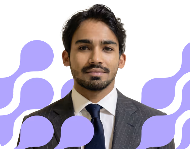 Portrait of a young man with dark hair and beard wearing a gray suit, white shirt, and dark blue tie in front of a background with purple abstract shapes.