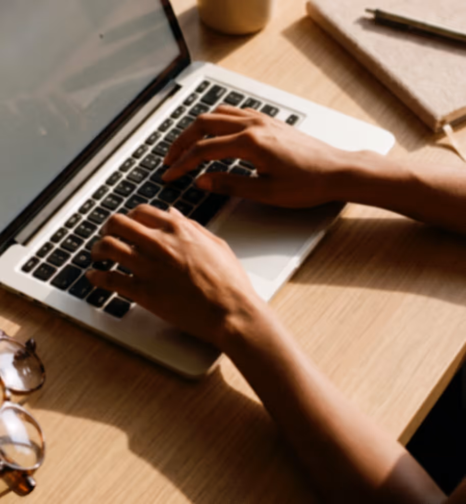 Person typing on a laptop keyboard on a wooden desk with glasses and a notebook nearby.
