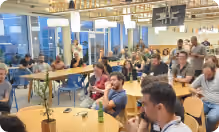 A group of people seated at tables watching a presentation or event in a bright room with large windows.