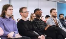 People sitting attentively in a room, facing forward, some taking notes.