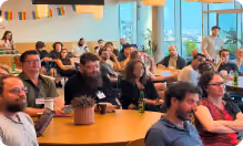 Group of people sitting and listening attentively in a bright room with large windows and colorful decorations.