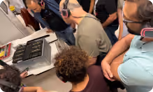 Group of people wearing headphones leaning over a table looking at an electronic device.