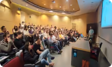 Audience seated in a lecture hall listening to a speaker at a podium with a projected presentation.
