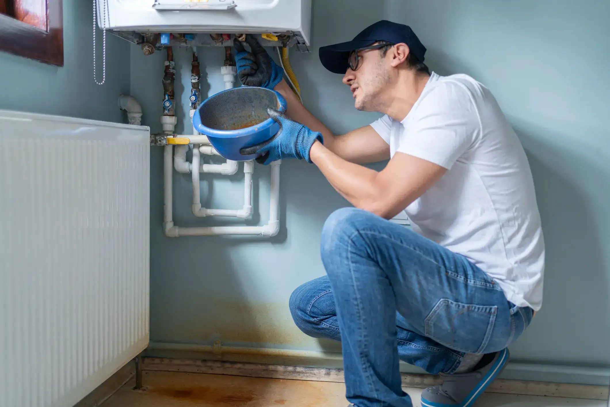 Plumber in blue cap and jeans inspecting pipes under a boiler with a blue bowl in hand
