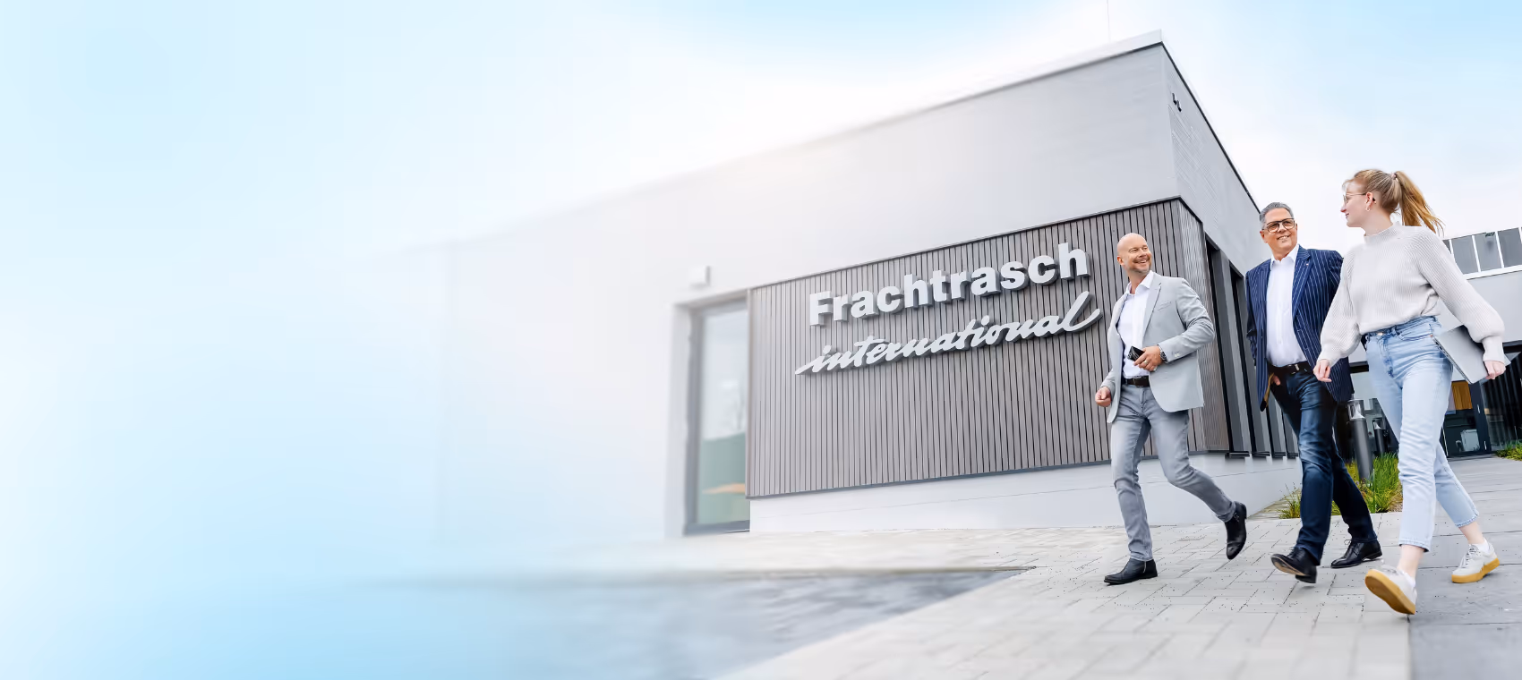 Drei Personen gehen lächelnd vor dem Firmengebäude von Frachtrasch International, moderne Unternehmensfassade mit Logo im Hintergrund.