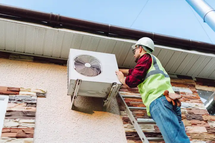 HVAC technician on a ladder installing a heat pump on the side of a house.