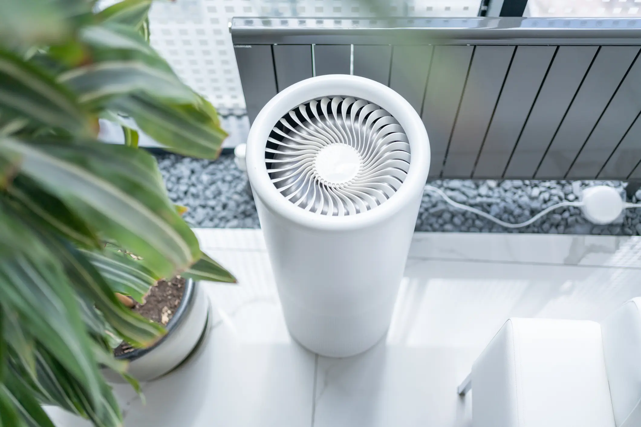 An overhead view of a white, cylindrical air purifier standing on a light-colored tiled floor. The unit features a distinctive circular top grille with a spiral fan pattern. A large potted plant is visible on the left, and a metal heating unit is in the background near a window and gray gravel.