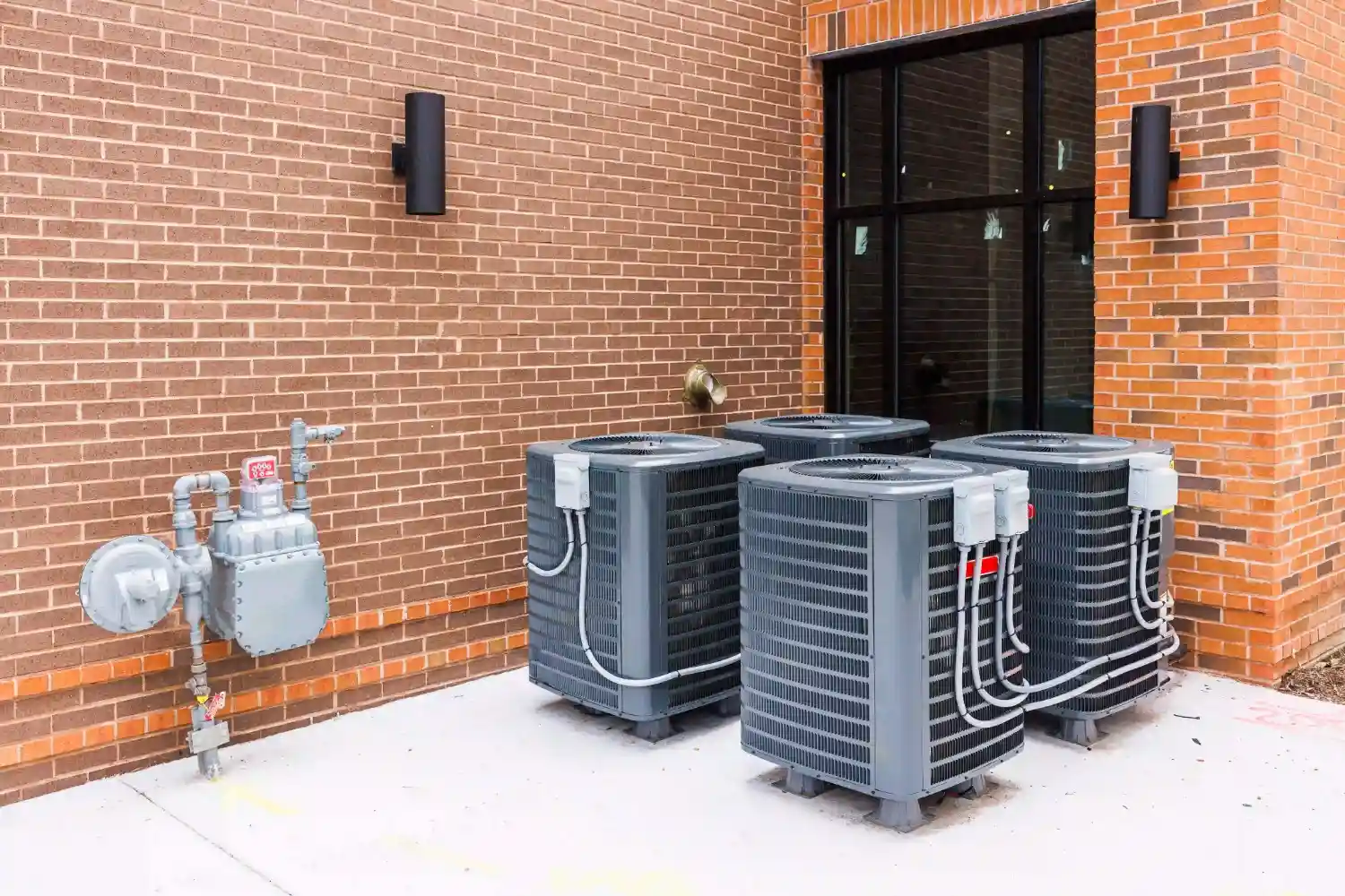  Three identical, gray outdoor HVAC units (likely air conditioners or heat pumps) are grouped together against a red brick commercial building. A gas meter and associated piping are visible on the left side, indicating the building uses both electricity and gas. The units are connected to the building via multiple white conduits and cables.