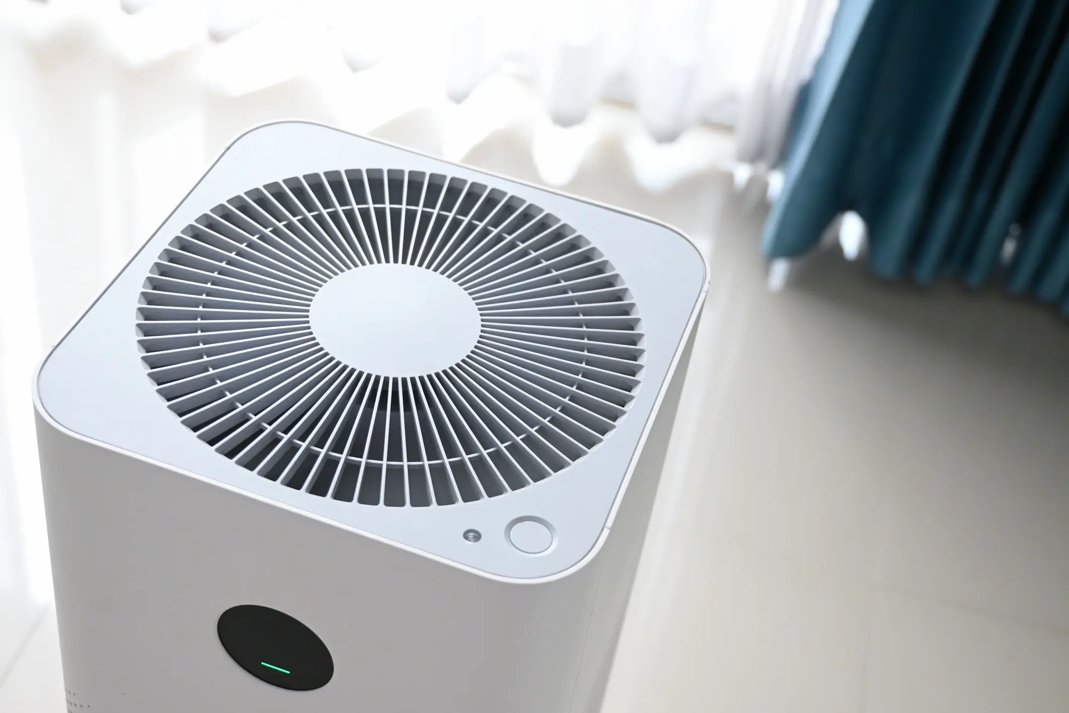 A close-up, high-angle view of the top of a white, modern air purifier. It features a large circular grille and a small power button/indicator light. The unit is sitting on a shiny floor near a window with sheer and blue curtains, suggesting it is operating in a bright, residential setting.