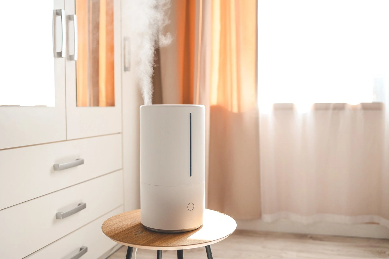 A modern, cylindrical, white humidifier is actively releasing a plume of steam or cool mist from its top. The unit, which has a vertical water level indicator line, sits on a small round wooden table. It is placed near a large, bright window with orange and white curtains and a white dresser/wardrobe.