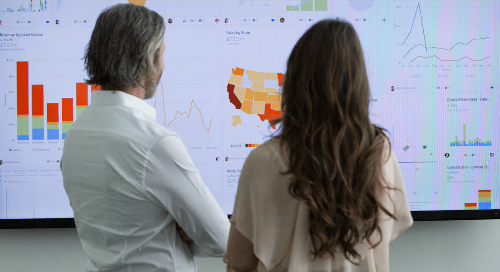 Two people standing and looking at a large screen displaying various business charts and graphs including bar charts and a colored map of the United States.