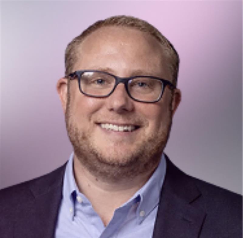 Smiling man wearing glasses, a light blue button-up shirt, and a dark blazer against a purple background.