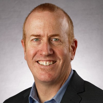 Middle-aged man with short reddish hair, wearing a dark blazer and blue collared shirt, smiling against a light gray background.