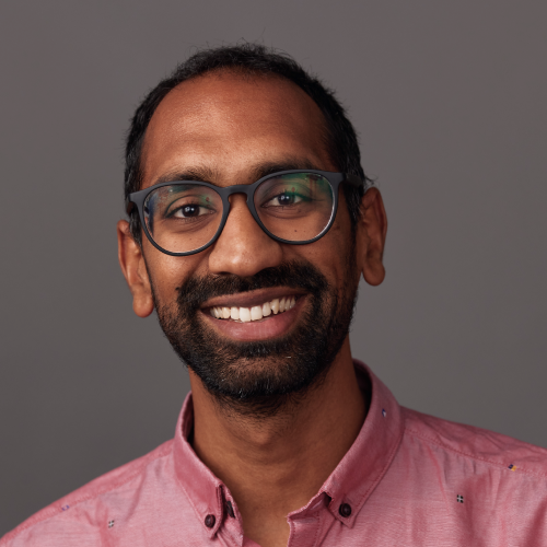 Smiling man with glasses and beard wearing a pink button-up shirt against a gray background.